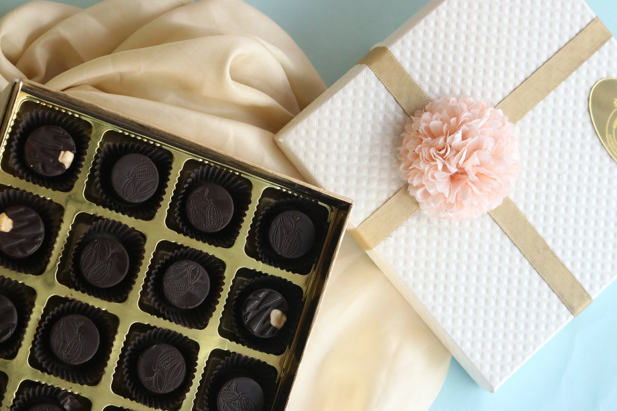 An open box of Dunkel Braun chocolates beside a white gift box tied with a gold ribbon and a peach flower